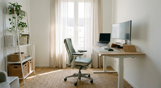A bright, ergonomically arranged home office in a modern rented flat, featuring a freestanding adjustable desk, monitor riser, and supportive chair bathed in natural window light.