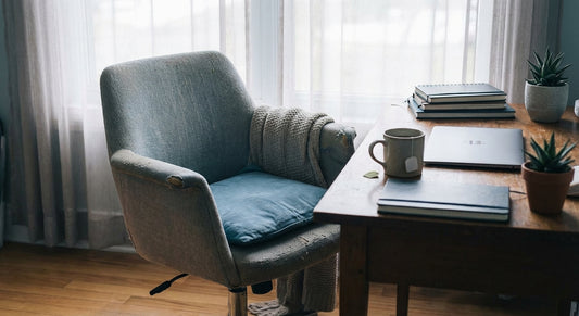 A dimly lit home office with an old, unsupportive office chair at a tidy desk beside a window, with a cup of tea and closed laptop suggesting interrupted work.